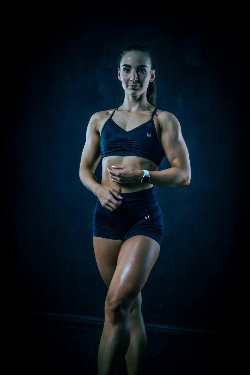 Dubi Anna sötétkék sportszettben, egyik kezét a hasán nyugtatva mosolyog a kamerába sötét stúdióban.