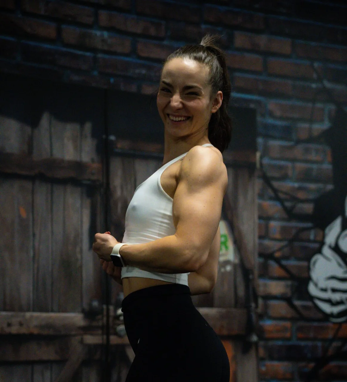 Dubi Anna nevetve mutatja a bicepszét egy téglafalas edzőteremben.